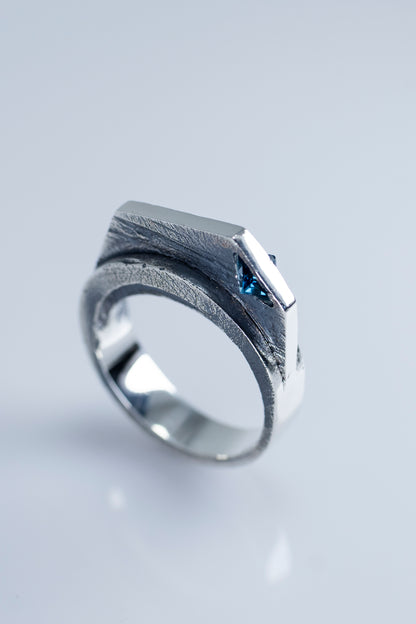 A modern silver statement ring with a geometric design, featuring a textured finish and a London Blue Topaz stone, photographed against a light background