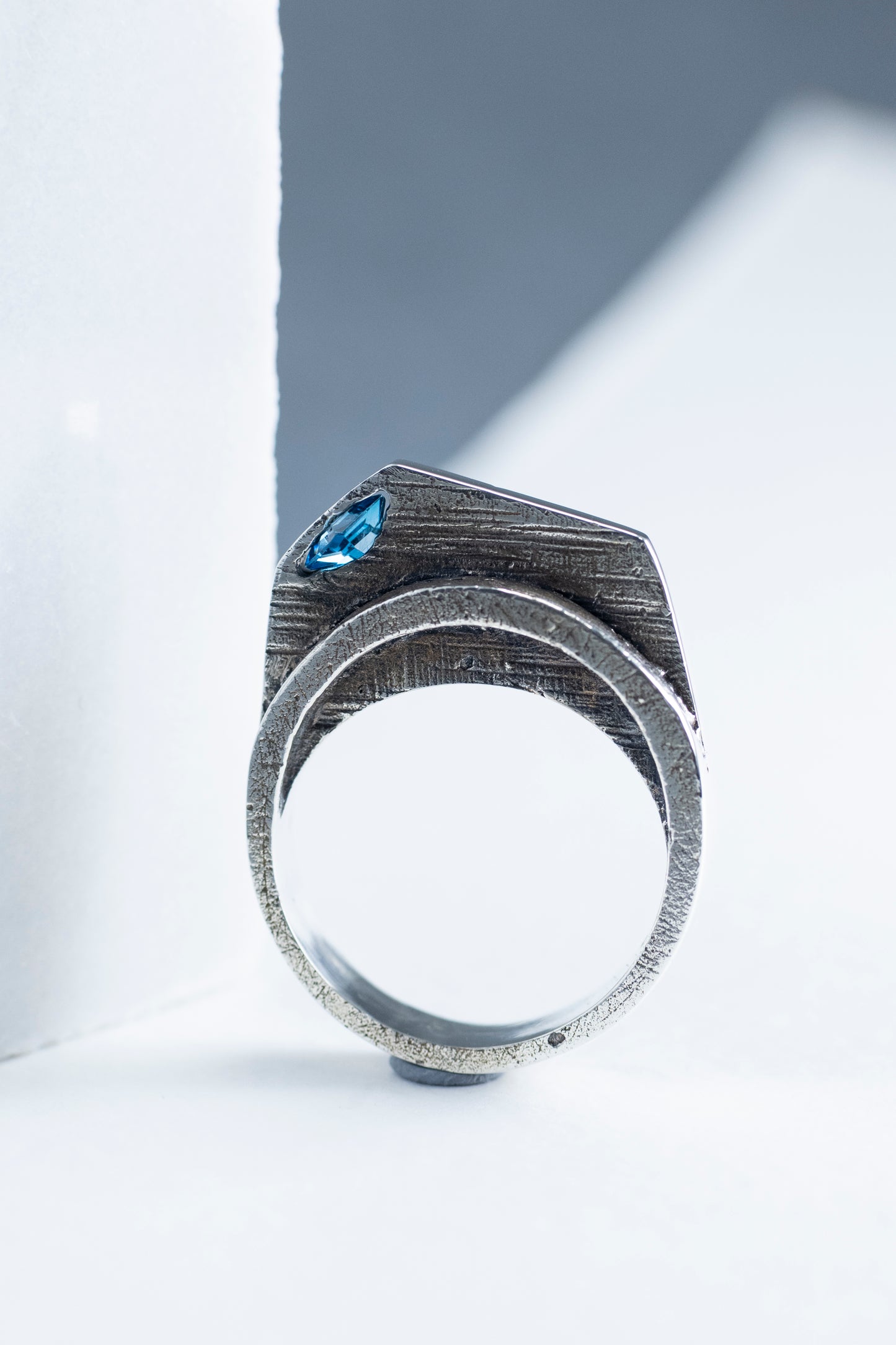 A close-up view of a silver statement ring showcasing its angular, textured design, with a London Blue Topaz stone set at an angle, highlighted against a soft, light background.